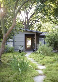 a small gray house surrounded by trees and grass with a path leading to the front door