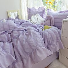 an unmade bed with purple comforter and white nightstands in front of a window