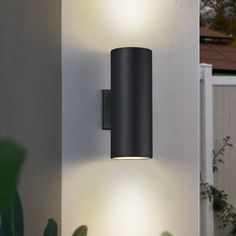 an outdoor wall light that is lit up on the side of a building with plants in front of it