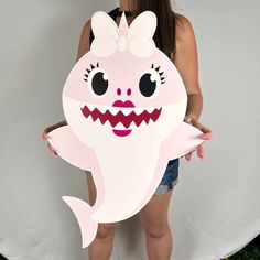 a woman holding up a cut out of a pink shark with big eyes and teeth