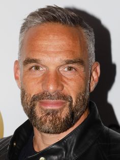 a man with grey hair and beard wearing a black leather jacket smiles at the camera