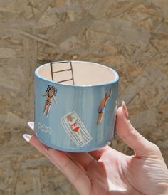 a hand holding a blue cup with people on the beach and lifeguards painted on it