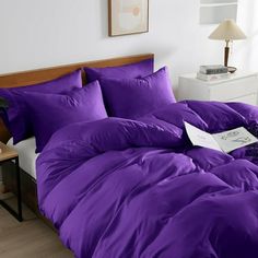 a bed with purple comforter and pillows in a white room next to a lamp