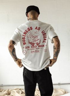 a man with tattoos standing in front of a white wall wearing a t - shirt that says business as usual ice cold beer