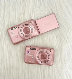 two pink digital cameras sitting on top of a white fur covered floor next to each other