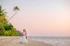 a bride and groom standing on the beach at sunset with palm trees in the background