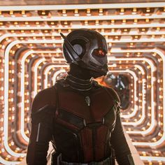 ant - man stands in front of an array of neon lights, with his helmet on