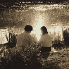 two people sitting on the shore of a lake in front of some grass and water