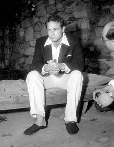 two men sitting on a bench in front of a stone wall, one wearing a suit and tie