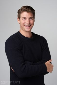 a man with his arms crossed smiling at the camera while wearing a black sweater and standing in front of a gray background