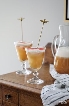 two glasses filled with drinks sitting on top of a wooden table next to a pitcher