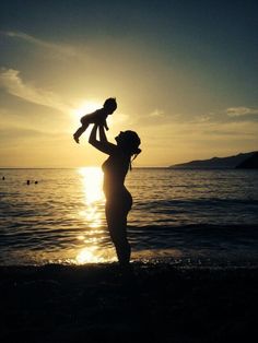 a woman holding a baby up in the air on top of a beach at sunset