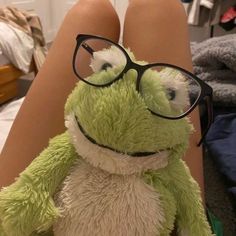 a stuffed frog wearing glasses sitting on top of a woman's legs in front of a bed