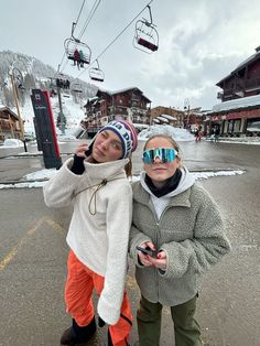 two young children standing next to each other in front of a ski lift with snow on the ground