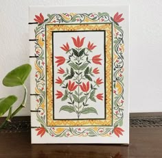 a white tile with red flowers and green leaves on the bottom is sitting next to a potted plant