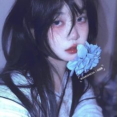 a woman with long black hair holding a blue flower in her mouth and looking at the camera