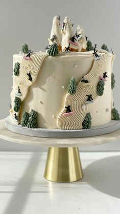 a white cake with trees and skiers on top is sitting on a gold pedestal