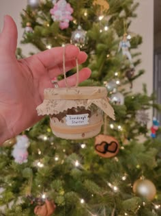 a hand holding a jar with some type of food in it next to a christmas tree