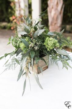 an arrangement of greenery in a wooden box on a white tablecloth with trees in the background