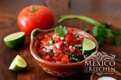 a bowl filled with salsa next to sliced tomatoes and avocado on a wooden table