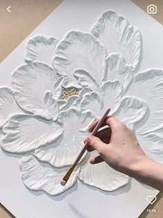 someone is painting a large flower with white paint on the paper and holding a brush in their hand