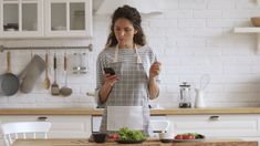 a woman standing in the kitchen looking at her cell phone while holding a knife and fork