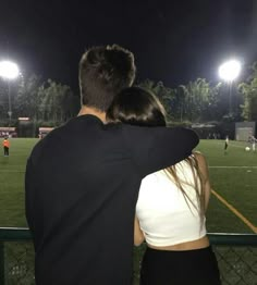 a man and woman sitting on the back of a bench watching a soccer game at night