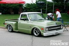 an old green truck parked in a parking lot with two men working on the hood