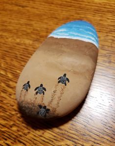 a close up of a painted rock on a wooden table with sand and sea turtles