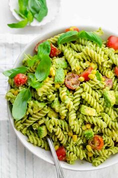 a white bowl filled with green pasta salad and topped with tomatoes, corn, spinach