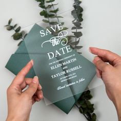 two hands holding an envelope with the word save the date printed on it, surrounded by greenery