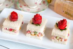 four small sandwiches with strawberries on them are sitting on a plate next to cups and saucers