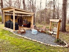 a small wooden shed in the middle of a forest with chairs and grilling area