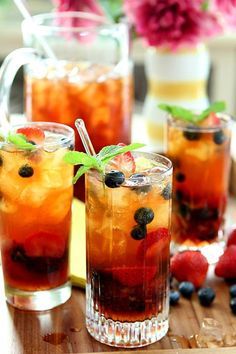 two glasses filled with fruit and ice on top of a wooden table next to flowers