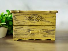 a yellow wooden box with a flower on the front and bottom, sitting next to a potted plant