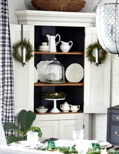 a white china cabinet filled with dishes and greenery