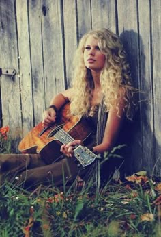 a woman with long blonde hair sitting on the ground holding an acoustic guitar in front of a wooden wall