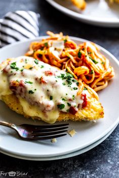 a white plate topped with chicken parmesan next to pasta and a fork on a table