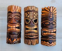 three wooden tiki masks are lined up against a white wall, each with different designs on their faces