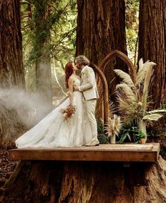 a bride and groom standing on a wooden platform in the woods surrounded by tall trees