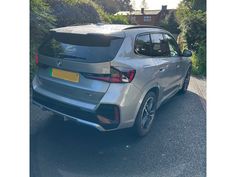 the rear end of a silver bmw suv parked in front of some bushes and trees