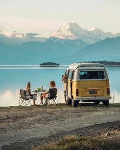 three people sitting at a table in front of a van on the shore of a lake