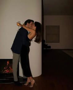 a man and woman kissing in front of a fire place with their arms around each other