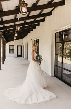 a woman in a wedding dress holding a bouquet standing on a porch with lights hanging from the ceiling