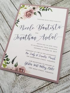 an elegant wedding card with pink flowers and greenery on the front, is displayed against a white wooden background