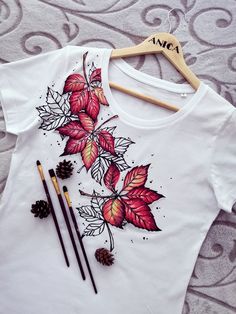 a white t - shirt with red leaves and pine cones on it, next to two pencils