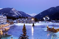 the town is lit up at night with christmas lights on trees and snow covered mountains in the background