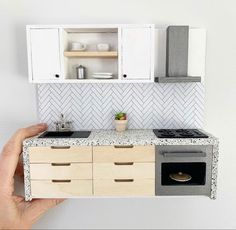 a hand holding a model of a kitchen with white cabinets and marble counter tops,