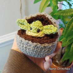 a crocheted baby yoda in a pot with a green plant behind it