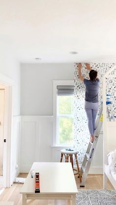 a woman standing on a ladder painting the wall in her living room with white walls